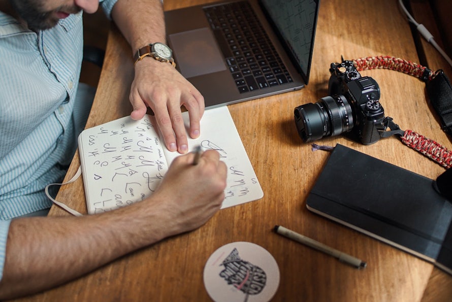 Man sketching in notepad with laptop and SLR camera in shot on wooden desk